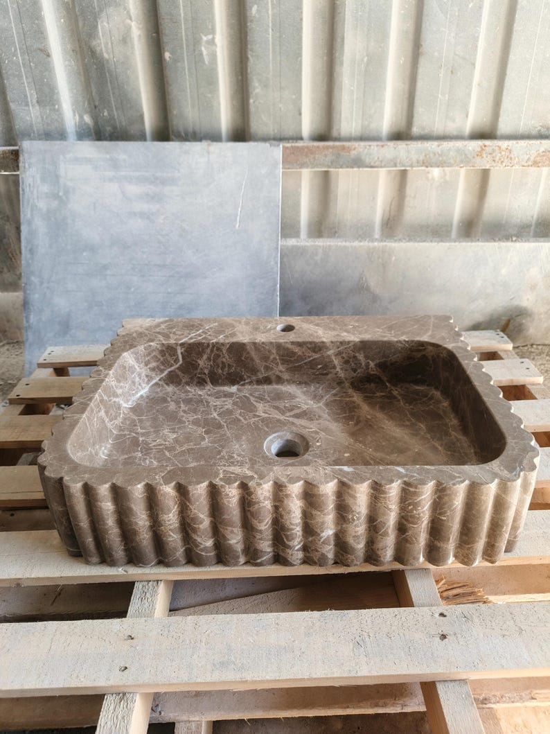 May include: A rectangular brown marble sink with a scalloped edge. The sink has a central drain and a faucet hole. The marble has a natural veining pattern. The sink is displayed on a wooden pallet.