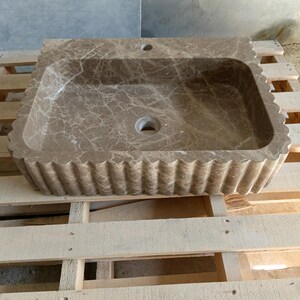 May include: A rectangular, brown marble sink with a scalloped edge. The sink has a rectangular basin with a drain hole and a faucet hole. The marble has a natural veining pattern. The sink is displayed on a wooden pallet.