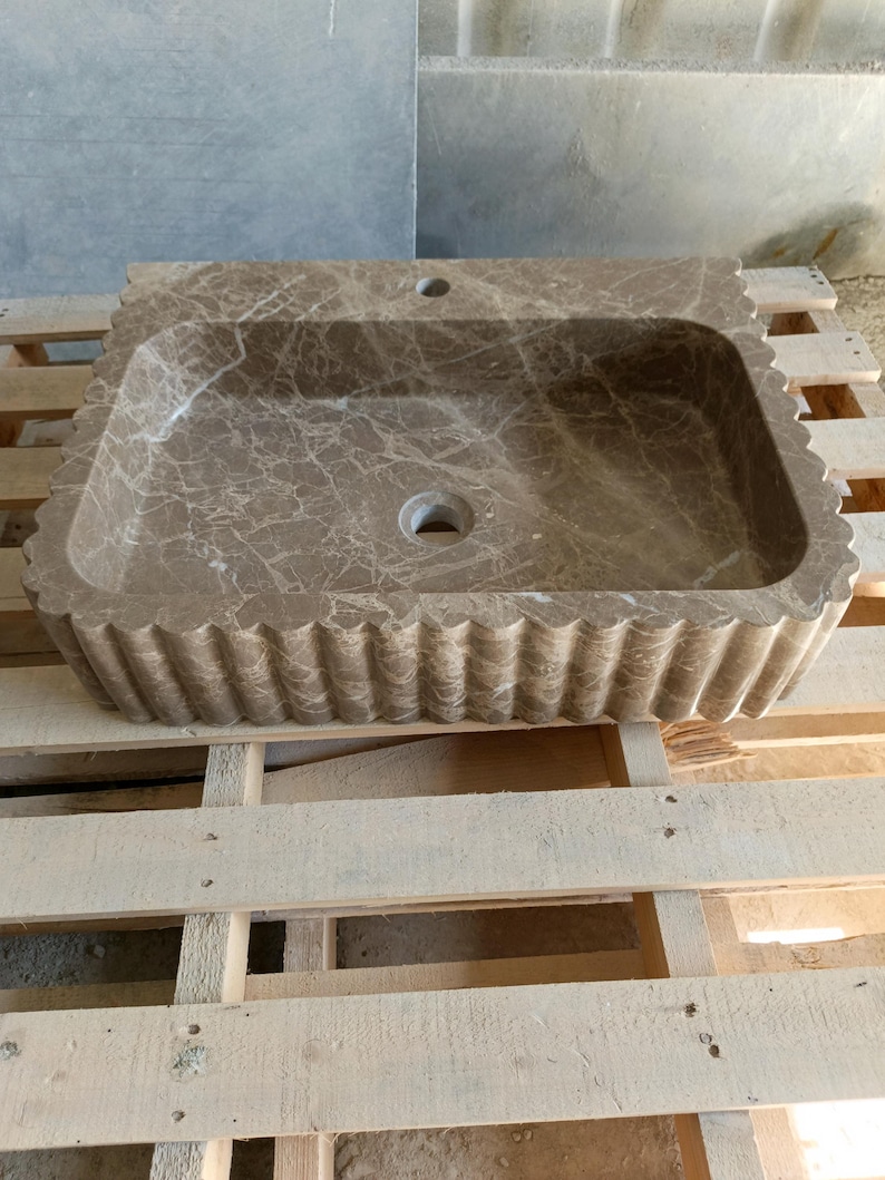 May include: A rectangular, brown marble sink with a scalloped edge. The sink has a rectangular basin with a drain hole and a faucet hole. The marble has a natural veining pattern. The sink is displayed on a wooden pallet.