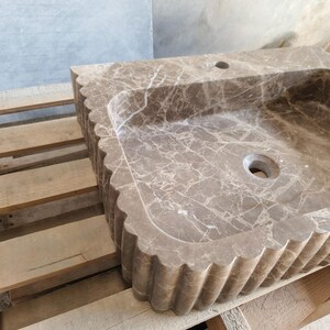 May include: A rectangular, brown marble sink with a scalloped edge. The sink has a basin with a drain hole and a faucet hole. The marble has a natural veining pattern. The sink is sitting on a wooden pallet.