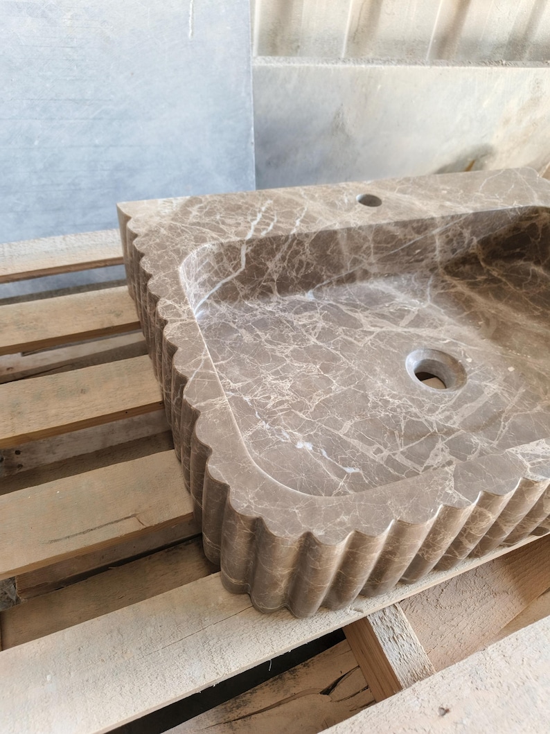 May include: A rectangular, brown marble sink with a scalloped edge. The sink has a basin with a drain hole and a faucet hole. The marble has a natural veining pattern. The sink is sitting on a wooden pallet.