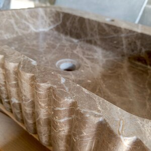 May include: A rectangular, brown marble sink with a decorative, ridged edge. The sink has a central drain hole and a natural veining pattern throughout the stone. The overall design is a blend of functionality and aesthetic appeal.
