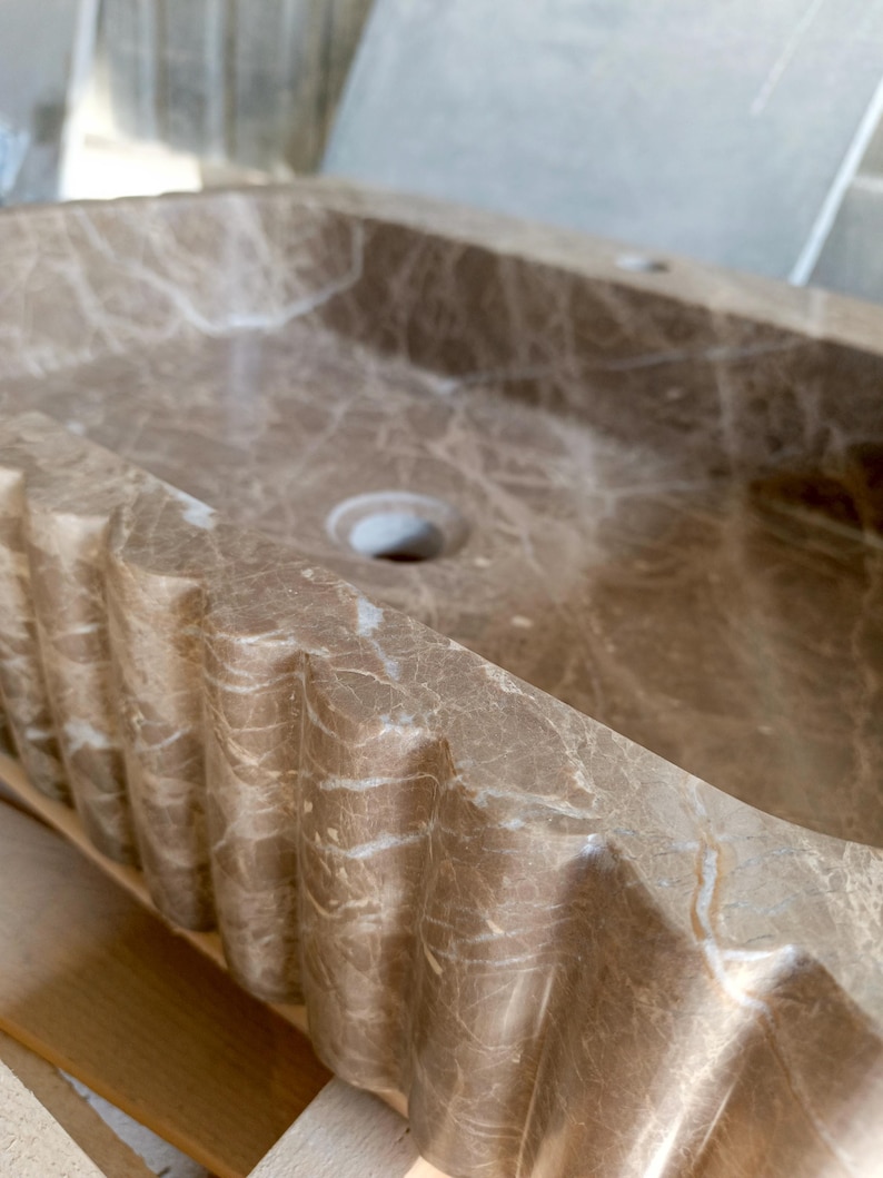 May include: A rectangular, brown marble sink with a decorative, ridged edge. The sink has a central drain hole and a natural veining pattern throughout the stone. The overall design is a blend of functionality and aesthetic appeal.