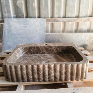 May include: A rectangular brown marble sink with a scalloped edge design. The sink has a natural veining pattern and is set on a wooden pallet. The sink is designed for bathroom use.