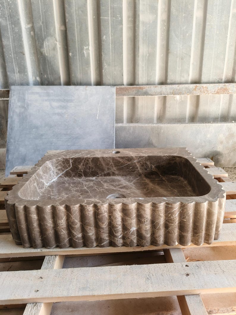 May include: A rectangular brown marble sink with a scalloped edge design. The sink has a natural veining pattern and is set on a wooden pallet. The sink is designed for bathroom use.