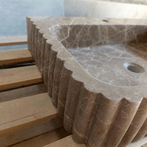 May include: A brown marble sink with a scalloped edge. The sink has a rectangular basin with a circular drain. The marble has a natural veining pattern. The sink is set on a wooden pallet.