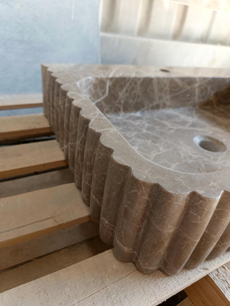 May include: A brown marble sink with a scalloped edge. The sink has a rectangular basin with a circular drain. The marble has a natural veining pattern. The sink is set on a wooden pallet.