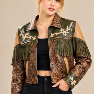 May include: A cropped jacket featuring a brown snakeskin pattern, olive green leather panels on the shoulders and cuffs, and long olive green fringe. The jacket includes cream-colored floral embroidery on the chest and cuffs, a collar, and a front zipper.