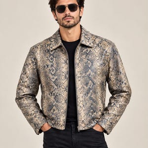 Peut inclure: Une veste à manches longues avec un motif peau de serpent beige et gris foncé, dotée d'un col classique et d'une fermeture éclair frontale. La veste est portée sur un t-shirt noir uni avec un pantalon noir et des lunettes de soleil aviateur.