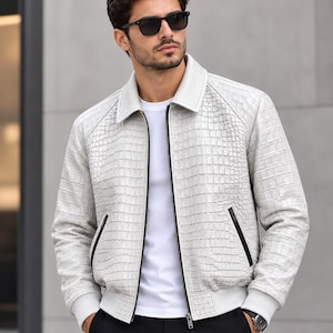 Peut inclure: Un blouson bomber en cuir blanc avec un motif texturé en peau de crocodile, un col classique, des manches longues à poignets côtelés et deux poches latérales à fermetures éclair noires. Porté sur un t-shirt blanc uni et un pantalon noir.