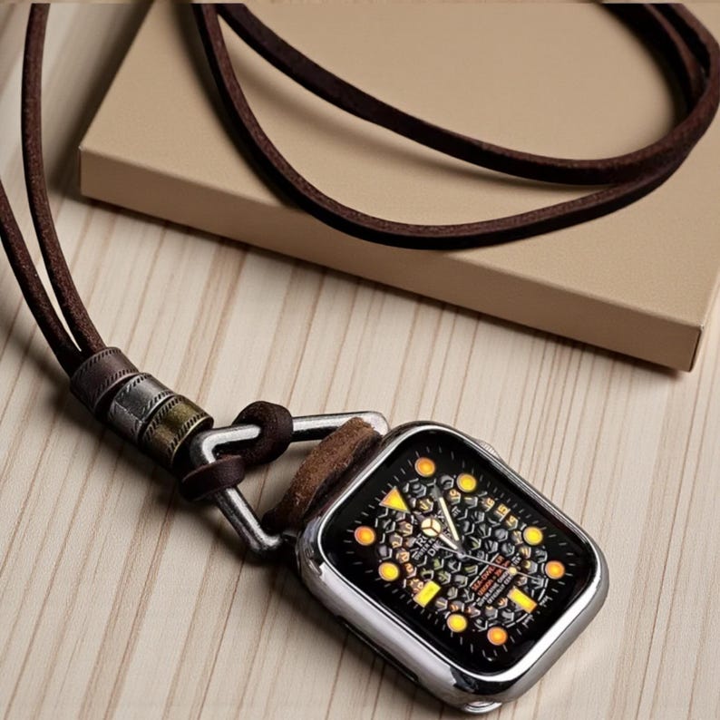 May include: A silver smartwatch case with a black face and yellow accents is attached to a brown leather cord necklace. The necklace is displayed on a light wooden surface, with a brown box in the background.