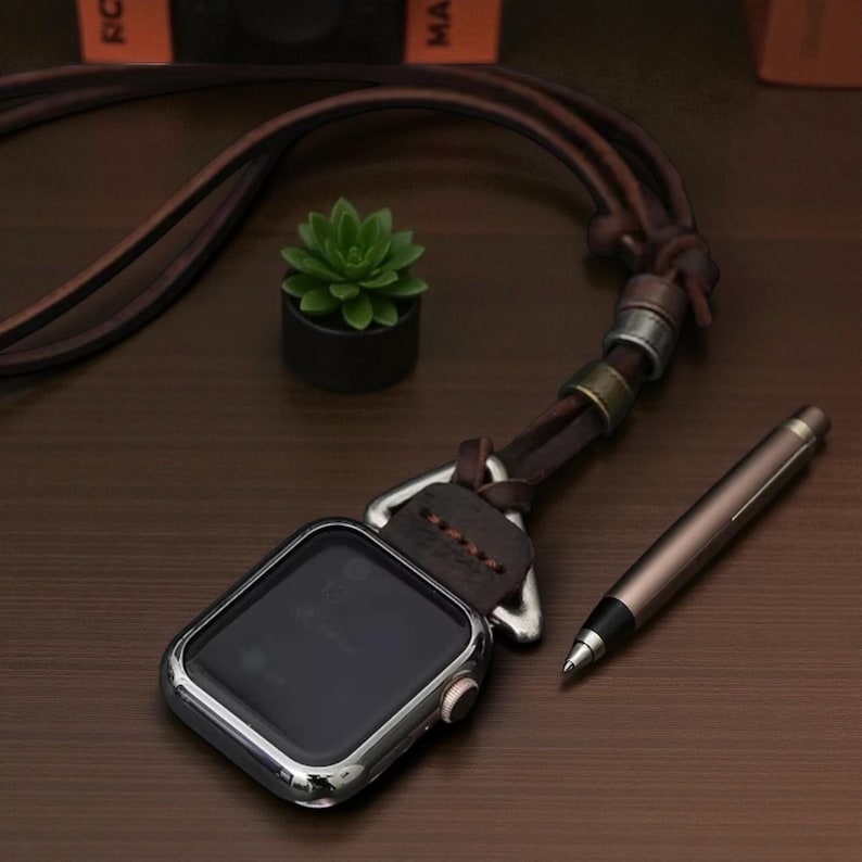 May include: A silver smartwatch with a dark screen, attached to a brown leather strap. A brown pen and a small succulent plant in a black pot are also on the dark wooden surface.