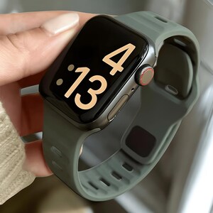 May include: A smartwatch with a black screen displaying the time as 13:04. The watch has a gray metal case and a green silicone band. The watch is being held by a person.