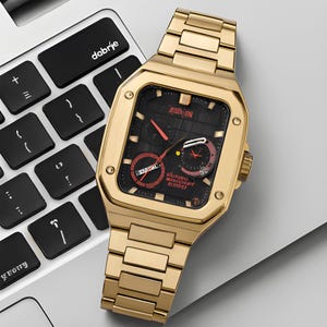 Può includere: Orologio da polso color oro con quadrante nero e dettagli rossi. L'orologio ha una cassa quadrata e un bracciale a maglie. Il quadrante mostra più quadranti e la data. L'orologio è su una tastiera di un laptop.