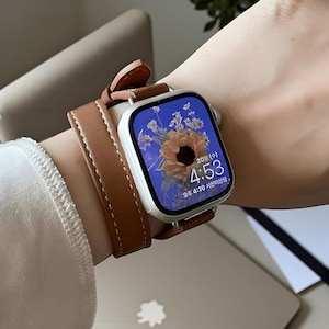 May include: A smartwatch with a light brown leather strap is worn on a wrist. The watch face shows a floral design and the time 4:53. The strap is secured with a buckle and has a stitched design.