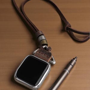 May include: A silver-framed smartwatch with a black screen, attached to a brown leather strap with metal accents. A matching pen lies nearby on a wooden surface. The watch strap has a loop for wearing around the neck.