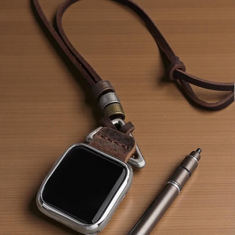 May include: A silver-framed smartwatch with a black screen, attached to a brown leather strap with metal accents. A matching pen lies nearby on a wooden surface. The watch strap has a loop for wearing around the neck.