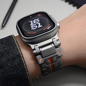 Könnte beinhalten: Graue Smartwatch mit digitaler Anzeige, die die Uhrzeit 16:51 und einen Batteriestand von 98 % anzeigt. Die Uhr hat ein graues Metallarmband mit orangefarbenen Akzenten. Die Uhr befindet sich an einem Handgelenk.