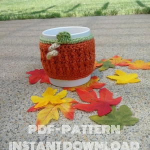 May include: Crocheted pumpkin-shaped coffee cup cosy in orange and green with a white ceramic mug. The cosy is surrounded by autumn leaves.