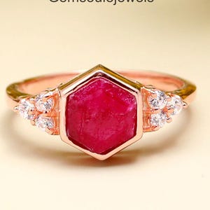 May include: Rose gold ring with a hexagonal, deep red gemstone. The ring has small, clear accent stones either side of the central stone. The text "Gemsoulejewels" is visible. A piece of jewellery.