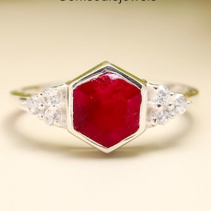 May include: A silver ring with a striking hexagonal, deep red gemstone at its centre. Either side of the main stone are clusters of smaller, clear gemstones. The ring is displayed against a soft, neutral background, with the text "Gemsoulejewels" visible.