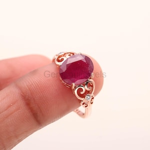 May include: Rose gold ring featuring a large, oval-cut ruby gemstone. The ring has a decorative filigree design on the band and small accent stones. The ruby is a deep, rich red color.