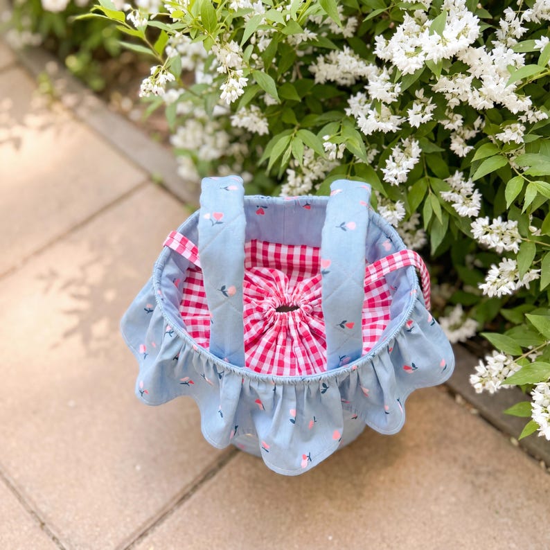 K&ouml;nnte beinhalten: Ein hellblauer Stoffkorb mit ger&uuml;schtem Rand und rosa Blumenmotiven. Im Inneren befindet sich ein rot-wei&szlig; karierter Kordelzugbeutel. Der Korb hat blau gesteppte Griffe und eine rosa Vichy-Verzierung.