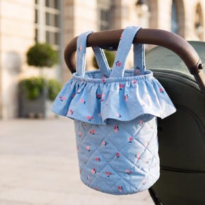 K&ouml;nnte beinhalten: Hellblau gesteppter Kinderwagen-Organizer mit einem Blumenmuster aus rosa Bl&uuml;ten. Der Organizer hat eine R&uuml;schenoberseite und Tr&auml;ger, die an einem braunen Kinderwagengriff befestigt sind. Der Hintergrund ist unscharf.