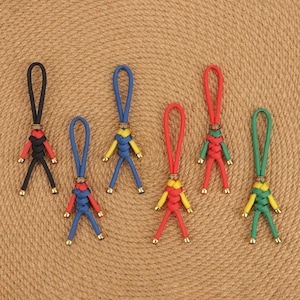 Puede incluir: Seis llaveros hechos a mano con diseño de figura humana. Cada llavero presenta un lazo, un nudo decorativo y un cuerpo con piernas. Los llaveros son en negro y rojo, azul y amarillo, rojo y amarillo, rojo y verde, y verde y amarillo.