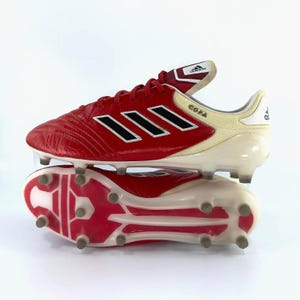 May include: Pair of red and white Adidas Copa soccer cleats. The cleats feature the Adidas logo and three black stripes on the side. The word "COPA" is printed on the side of the shoe. The cleats are designed for use on a soccer field.