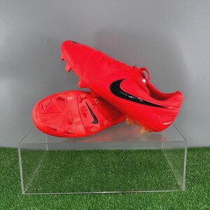 Op de afbeelding: Felrode voetbalschoenen met zwarte Nike swoosh-logo's. De schoenen worden gepresenteerd op een transparante acrylstandaard. De schoenen hebben meerdere noppen op de zolen.