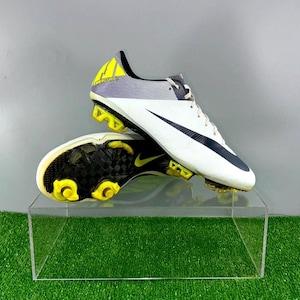 May include: Pair of white and black football boots with yellow detailing. The boots have a black swoosh logo and yellow studs on the sole. The heel area features yellow and grey detailing.