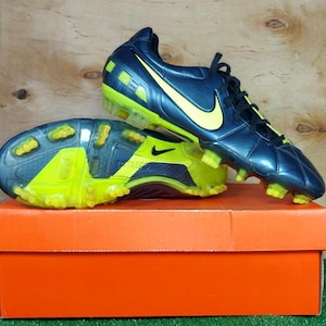 Könnte beinhalten: Ein Paar marineblaue und gelbe Nike-Fußballschuhe mit gelbem Nike-Swoosh-Logo. Die Schuhe stehen auf einer orangefarbenen Box.