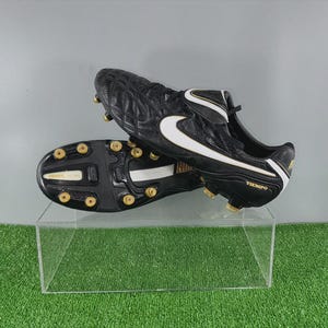 Könnte beinhalten: Schwarz-weiße Fußballschuhe mit goldenen Akzenten. Die Schuhe haben ein rautenförmiges Muster auf dem Obermaterial und ein großes weißes Swoosh-Logo. Die Sohle hat goldene Stollen. Das Wort "Tiempo" ist auf der Seite sichtbar.