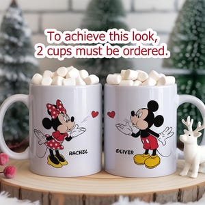 K&ouml;nnte beinhalten: Zwei wei&szlig;e Keramikbecher gef&uuml;llt mit Marshmallows, mit Minnie- und Mickey-Mouse-Designs. Auf einem Becher steht "Rachel" und auf dem anderen "Oliver". Der Text "To achieve this look, 2 cups must be ordered" ist ebenfalls sichtbar.