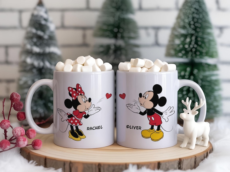Two white ceramic mugs filled with marshmallows, featuring Mickey and Minnie Mouse designs with a heart graphic.  Minnie wears a red polka-dot dress and bow, Mickey wears red shorts. Each mug has a name on it.