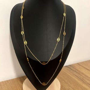 Könnte beinhalten: Eine goldfarbene Halskette mit einem doppelreihigen Design. Die Halskette hat ovale Glieder, die gleichmäßig über die zarte Kette verteilt sind. Der Schmuck wird auf einer schwarzen Mannequin auf einer Holzoberfläche präsentiert.
