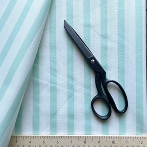 May include: A white fabric with light green stripes, folded over itself. A pair of black fabric scissors are lying on top of the fabric. A ruler with inch markings is visible at the bottom of the image.