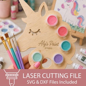 May include: A light-coloured wooden paint palette shaped like a unicorn head with a cutout eye and the text Aly's Paint STUDIO. It features five circular cutouts holding small paint pots in pink, red, teal, light green, and purple, with four paintbrushes.
