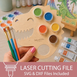 May include: A light-coloured wooden paint palette shaped like a dinosaur head with six circular cutouts holding small paint pots in green, blue, orange, yellow, and brown. The palette features the text Smiles and Colors STUDIO and three paintbrushes.