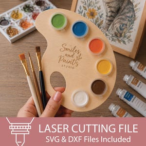 Könnte beinhalten: Eine hellfarbene Holz-Malpalette mit der lasergravierten Aufschrift Smiles and Paints STUDIO. Die Palette enthält sechs kreisförmige Aussparungen für kleine Farbtöpfe in Grün, Blau, Orange, Gelb, Braun und Weiß sowie drei verschiedene Pinsel.