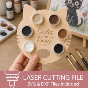 May include: A light-coloured wooden paint palette shaped like a cat head with six circular cutouts holding small paint pots. The centre is engraved with the text Smiles and Paints STUDIO. The palette features a thumb hole at the bottom.