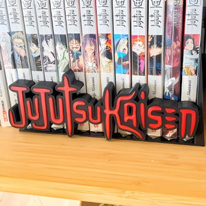 May include: A black and red "Jujutsu Kaisen" logo, likely a shelf decoration, stands in front of a row of manga books. The logo is 3D printed with a red outline and a black fill. The books have white spines with Japanese text.