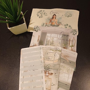 Könnte beinhalten: Ein Set von Yoga-Karten und einem passenden Beutel. Der Beutel zeigt eine Illustration einer meditierenden Frau mit dem Wort "Ommm" darüber. Die Karten haben Innenarchitektur-Illustrationen und Text. Eine kleine Topfpflanze ist ebenfalls vorhanden.