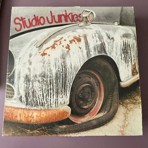 May include: A vintage-style image featuring a weathered car with the text "Studio Junkies" in red. The car's body shows signs of rust and wear, with a flat tire. The overall aesthetic is rustic and nostalgic.