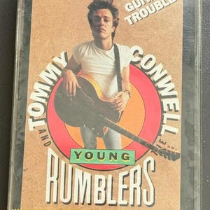 May include: A cassette tape featuring the album cover for "Guitar Trouble" by Tommy and the Young Rumblers. The cover art shows a young man holding a guitar, with text in white and green.