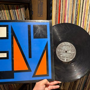 Może przedstawiać: Winyl Split Enz "True Colours". Okładka albumu ma niebieskie tło z pomarańczowymi i czarnymi geometrycznymi kształtami. Czarny winyl leży obok okładki. Etykieta jest widoczna.