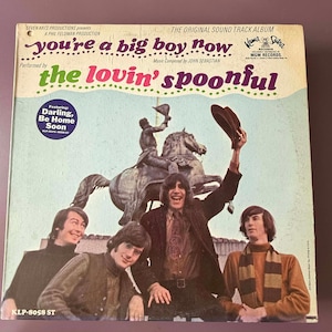 以下が含まれることがあります： The Lovin' Spoonfulの「You're a Big Boy Now」のヴィンテージビニールレコードアルバムのカバー。カバーには、背景に像のあるバンドの写真が掲載されています。アルバムタイトルとバンド名はカラフルなデザインです。