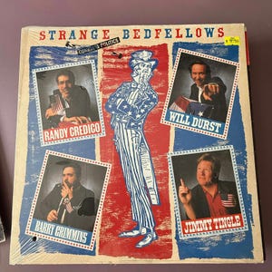 Può includere: Un album in vinile intitolato "Strange Bedfellows" con il sottotitolo "Comedy & Politics". La copertina presenta lo Zio Sam e ritratti di Randy Credico, Will Durst, Barry Crimmins e Jimmy Tingle. Lo sfondo è rosso, bianco e blu.