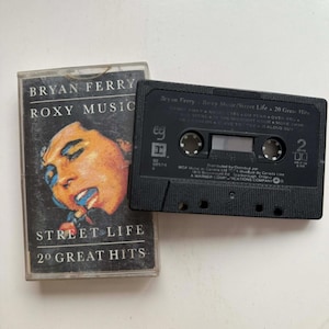May include: A vintage cassette tape of Bryan Ferry's "Roxy Music: Street Life - 20 Great Hits." The cassette case features a portrait of a person with orange and blue tones. The black cassette tape is next to the case.